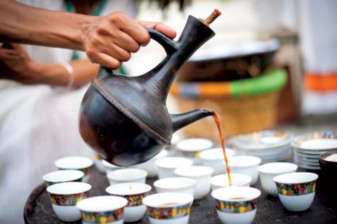 A hand pours coffee out of a tall brown ceramic pot into many small white cups
