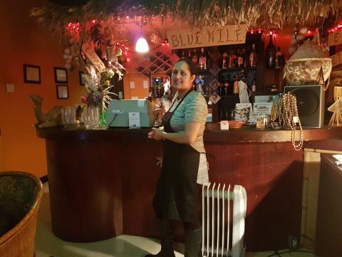 The restaurant owners wife Asmeret stands in front of the counter with the grass canopy and "Blue Nile" sign, a collection of traditional East African arts and artifacts decorates the counter