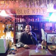 The owner of the restaurant Michael stands behind a counter decorated with a grass canopy and wooden sign that reads "Blue Nile"